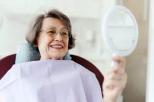 Senior woman smiling at herself in the mirror after her routine dental checkup.