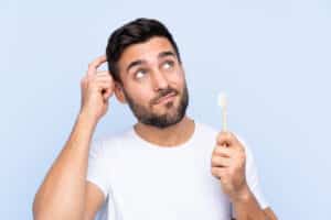 Man scratching head while holding a toothbrush, appearing unsure about when to brush his teeth.